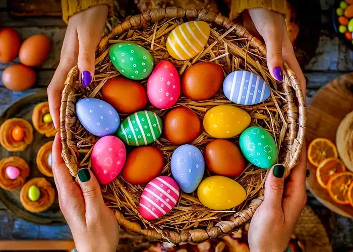 Hands holding a basket of brightly colored Easter eggs with patterns on a rustic wooden table